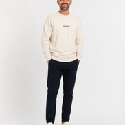 Man wearing a cream sweater with a logo and dark pants on a white background