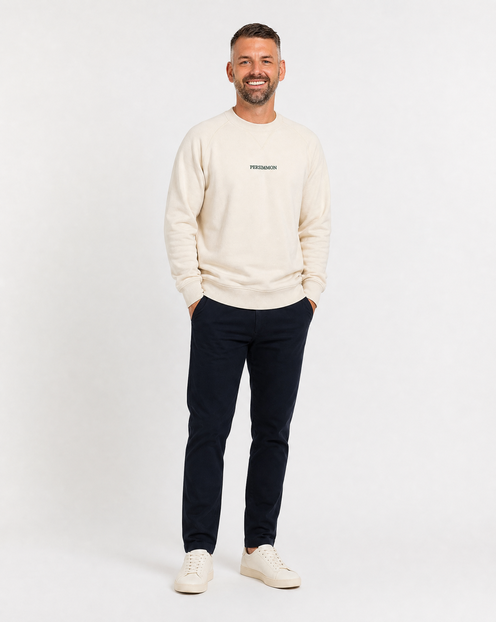 Man wearing a cream sweater with a logo and dark pants on a white background