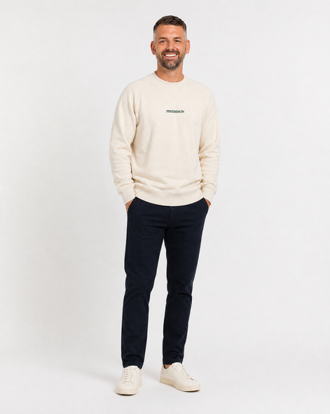Man wearing a cream sweater with a logo and dark pants on a white background