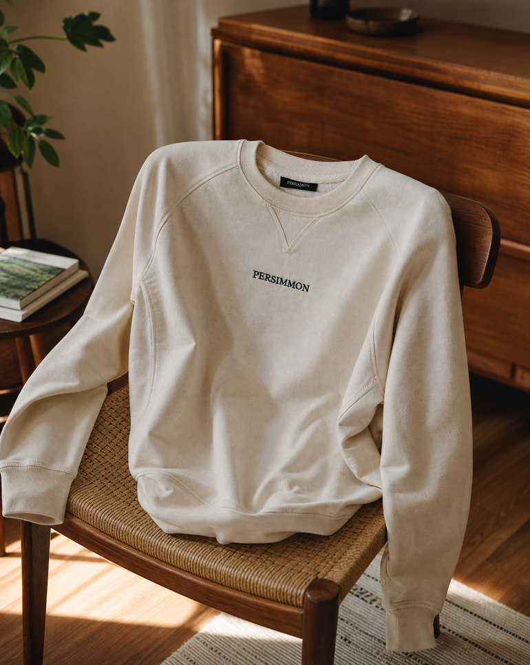 A cream crewneck sweatshirt with 'PERSIMMON' printed in dark green on the chest, draped over a woven-back wooden chair in a warm, sunlit room.