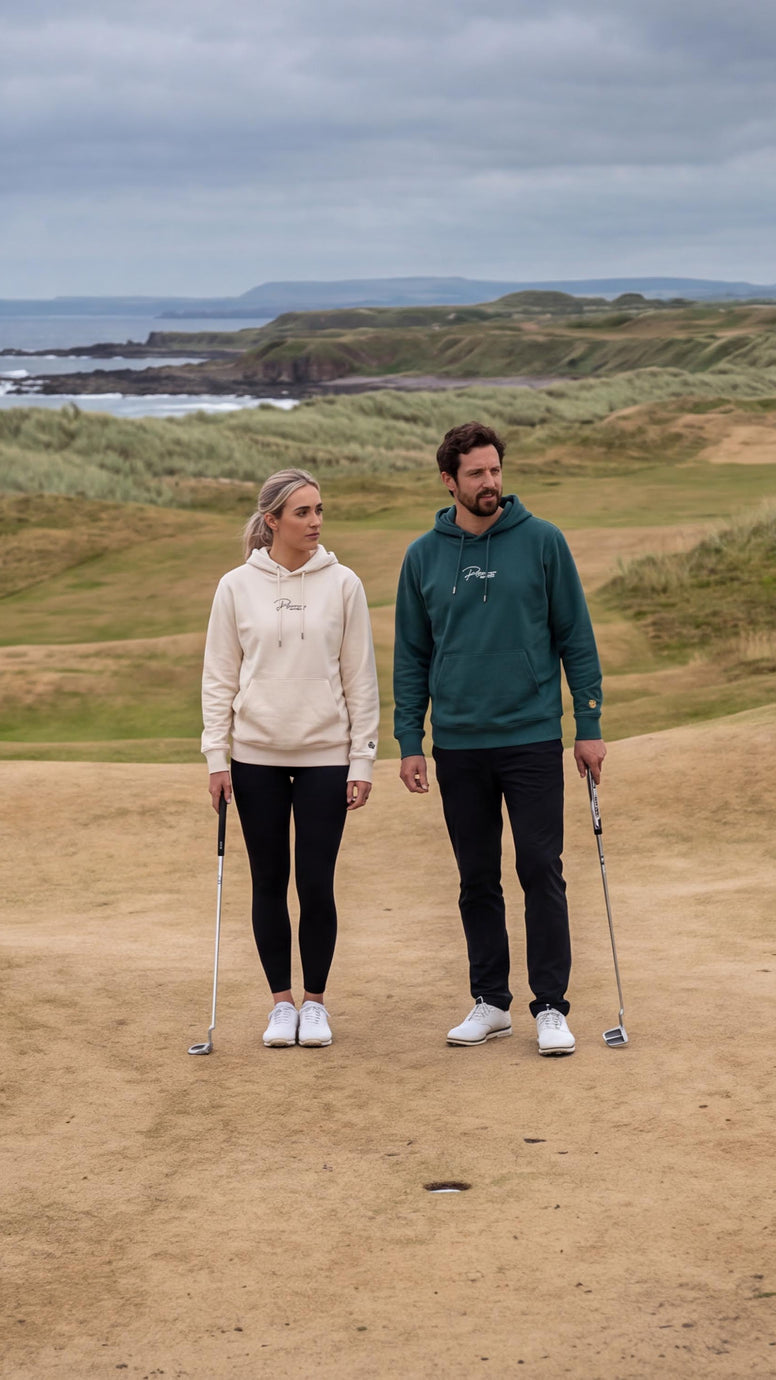 A man and a woman standing on a golf course green, each holding a golf club. The woman wears a cream hoodie and black pants, and the man wears a dark green hoodie and black pants.
