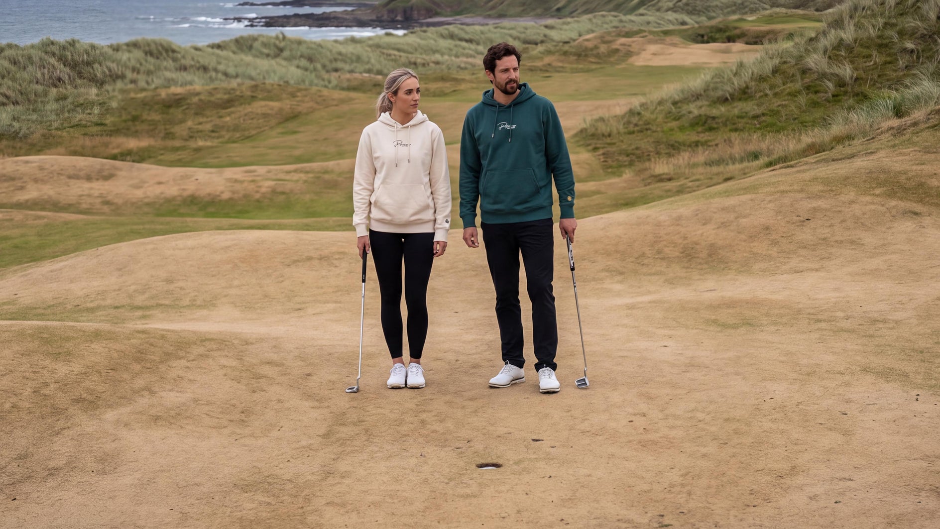Two people standing on a sand bunker at a coastal golf course, with grassy dunes, ocean, and an overcast sky in the background.