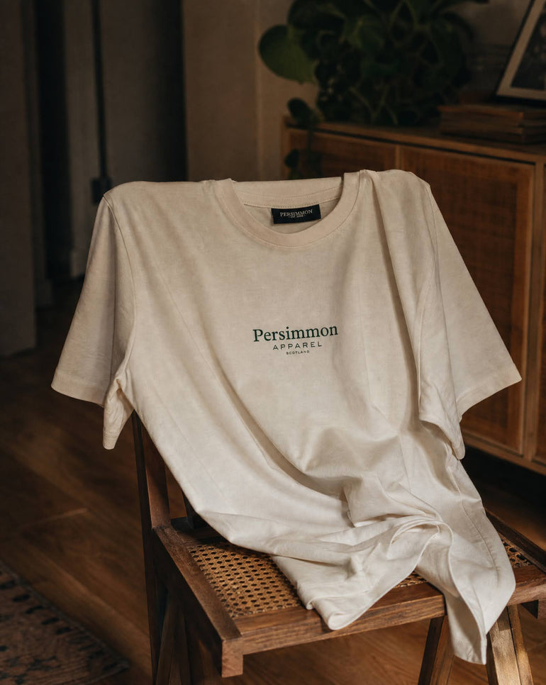 A cream short-sleeve t-shirt with 'Persimmon APPAREL' printed in dark geen on the chest, draped over a wooden chair in a warm, dimly lit indoor setting.