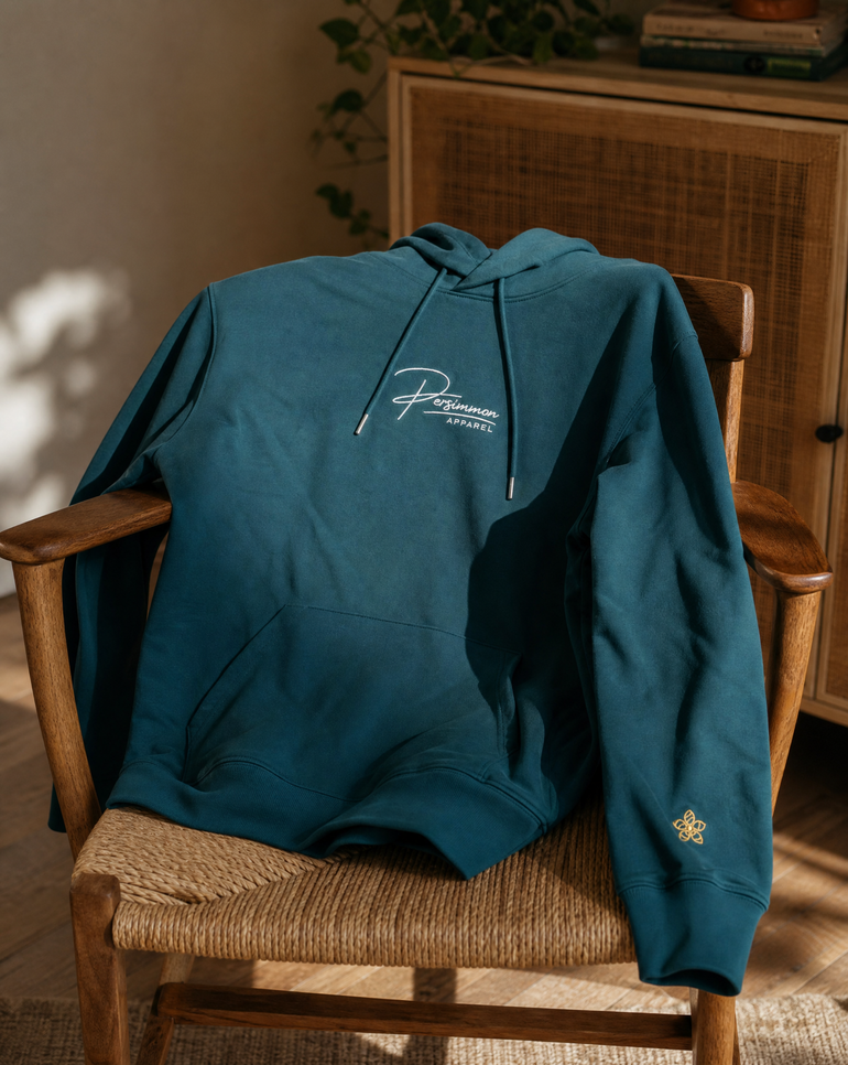 A green pullover hoodie with a white embroidered 'PERSIMMON' script logo on the chest and a small gold embroidered emblem on the sleeve, draped over a wooden chair in a warm, sunlit room.
