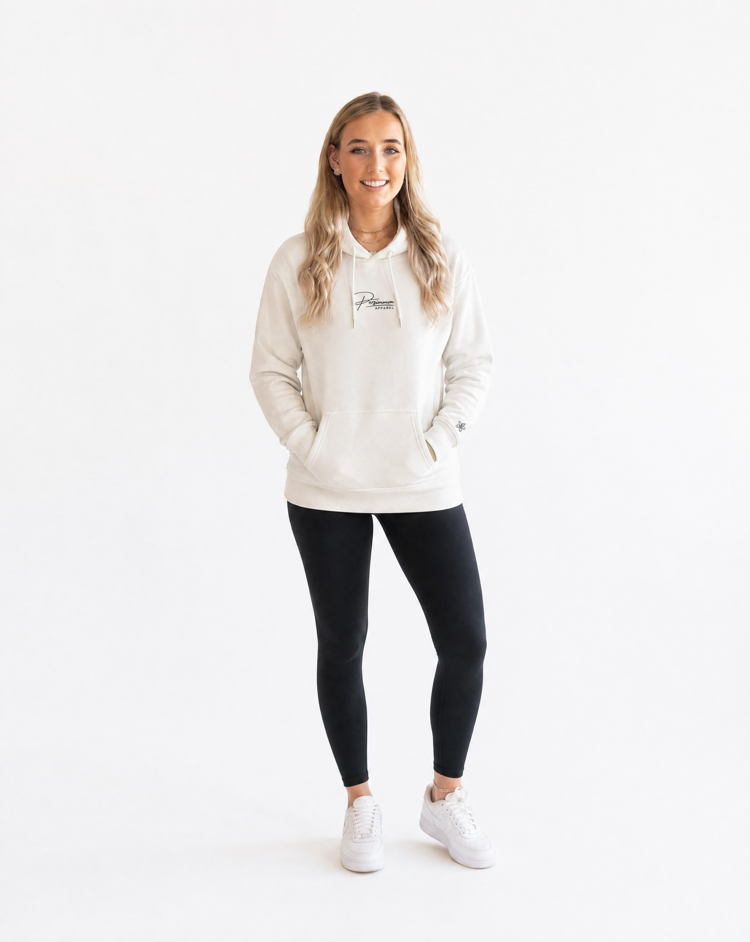 Woman wearing a cream hoodie with Persimmon logo, black leggings, and white trainers on a white background